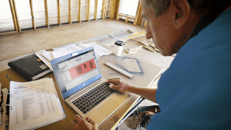 Small residential builder reviewing estimates and job costs on a laptop at a construction site to track pricing and margin during active work.