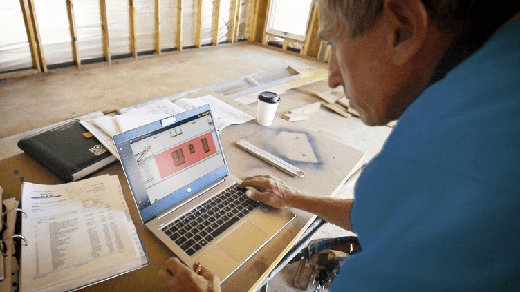 Residential builder reviewing construction plans and job data on a laptop at an active build site
